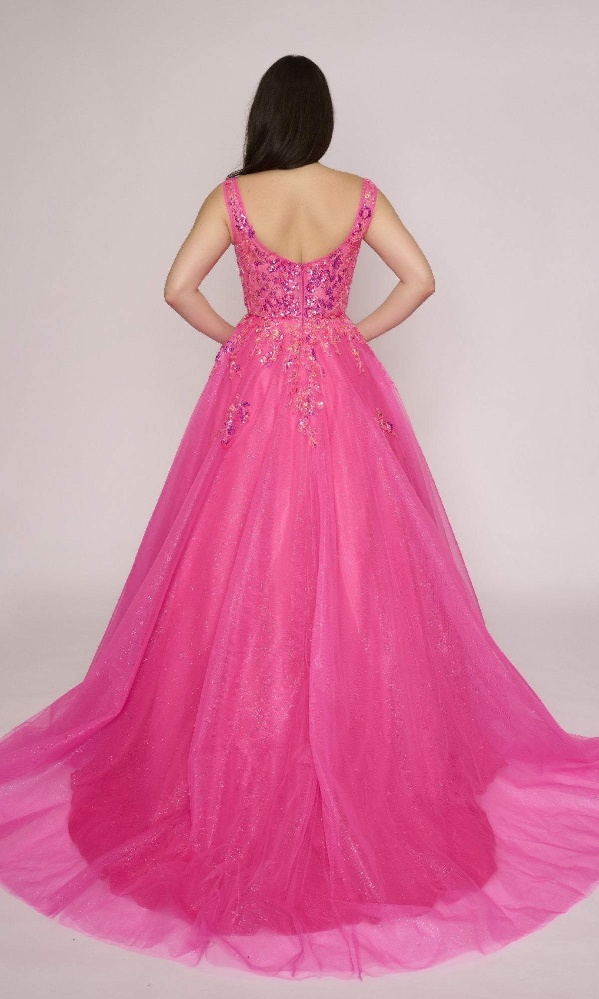 A woman facing away, wearing the Nina Canacci 3266 long prom dress in bright pink with floral embellishments and a sheer glitter tulle overlay.